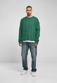 Green cable-knit sweater with ribbed cuffs, worn over a white shirt. Paired with faded blue jeans and white sneakers. Standing against a gray background.