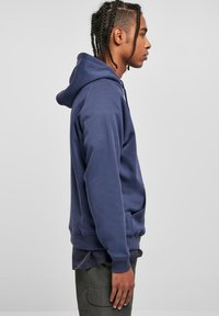 Sudadera azul marino con capucha ajustable, puños acanalados y bolsillo canguro, llevada sobre una camiseta oscura, mostrada de lado.