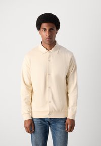 Camicia button-up di colore crema con colletto, realizzata in tessuto morbido. Presenta una vestibilità rilassata e polsini a costine, abbinata a jeans di denim blu.