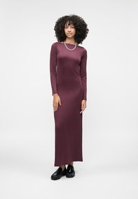 Duga rukava, bordo haljina s uskim krojem, izrađena od glatkog, rastezljivog materijala s ravnim rezom do članka. Uparena s crnim cipelama.