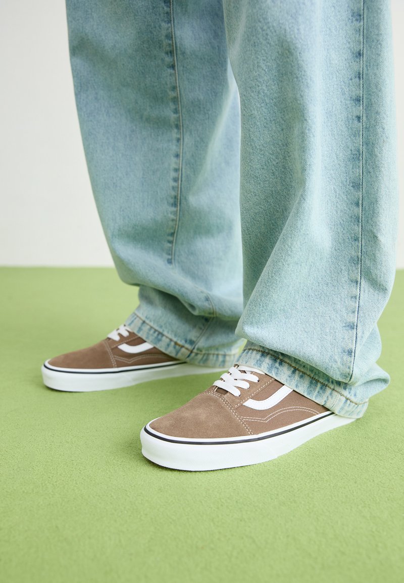 Baskets en daim marron avec des accents et des lacets blancs, associées à un jean baggy bleu clair, sur un fond vert.