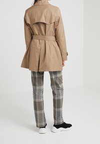 Kvinna iklädd en beige trenchcoat med bälte, rutig byxor och svarta sneakers med vita sulor, stående i ett vitt rum med ryggen mot betraktaren.