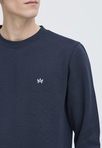 Mørkeblå sweatshirt med rund halsudskæring, ribbede manchetter og et lille hvidt krone-logo på brystet. Fremstillet af struktureret stof.