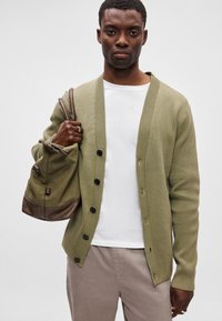 Olivovo zelený rebrovaný kardigán s čiernymi gombíkmi, nosený na bielom tričku. Model drží ramennú tašku s hnedými akcentmi, doplnenú béžovými nohavicami.