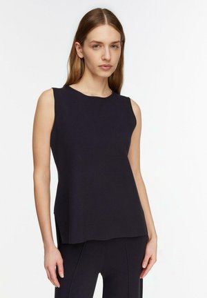 Młoda kobieta ubrana w czarny, luźny bezrękawnik i dopasowane czarne spodnie, stojąca na tle jednolitej białej ściany.