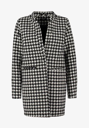 Houndstooth-takki mustassa ja valkoisessa, pitkät hihat, napittomat etuosat, yhdistettynä sinisiin farkkuihin ja mustaan kassi. Kiiltävät nilkkurit.