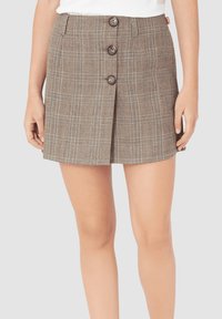 Mujer con una minifalda de cuadros marrón de botones en la parte delantera, con lazos para cinturón y una pequeña etiqueta roja en la cintura.