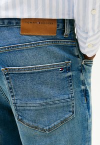 Jeans in denim blu con un lavaggio chiaro, caratterizzati da una patch in pelle con l'etichetta "TOMMY HILFIGER" e un piccolo logo sulla tasca posteriore.