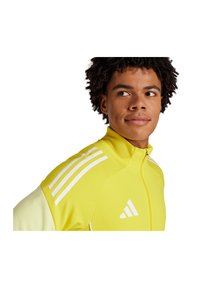 Gelbe Sportjacke mit weißen Akzenten und einem Drei-Streifen-Design auf den Schultern. Hergestellt aus glattem synthetischem Material mit hohem Kragen.