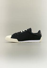 Μαύρο αθλητικό παπούτσι χαμηλού προφίλ με λευκή ελαστική σόλα, προστατευτικό μύτης και κορδόνια, εμφανιζόμενο από το πλάι σε απλό φόντο.