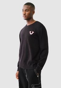 Schwarzer Pullover mit geripptem Saum und Bündchen, der ein pinkes Logo auf der Brust zeigt. Weicher Stoff mit einem klassischen Rundhalsausschnitt.