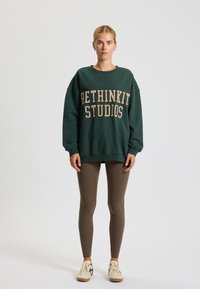 Felpa verde con "BETHINKIT STUDIOS" stampato in beige, abbinata a leggings marroni e sneakers crema con dettagli neri.
