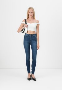 Top alb off-the-shoulder cu margini voluminoase, blugi skinny albaștri și pantofi cu vârf ascuțit negri. Modelul poartă o poșetă alb-negru.