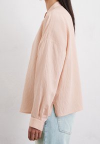 Chemise oversize rayée rose clair avec col montant, épaules tombantes et manches longues, associée à un jean bleu clair. Tissu texturé.