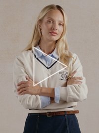 Kremowa bluza z dekoltem w serek, granatowym wykończeniem i logo; noszona na jasnoniebieskiej koszuli; zestawiona z ciemnymi jeansami i brązowym paskiem.