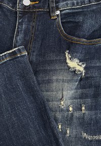 Donkerblauwe denims met gele stiksels, voorzak, knoop en versleten scheurdetails ter hoogte van de dij.