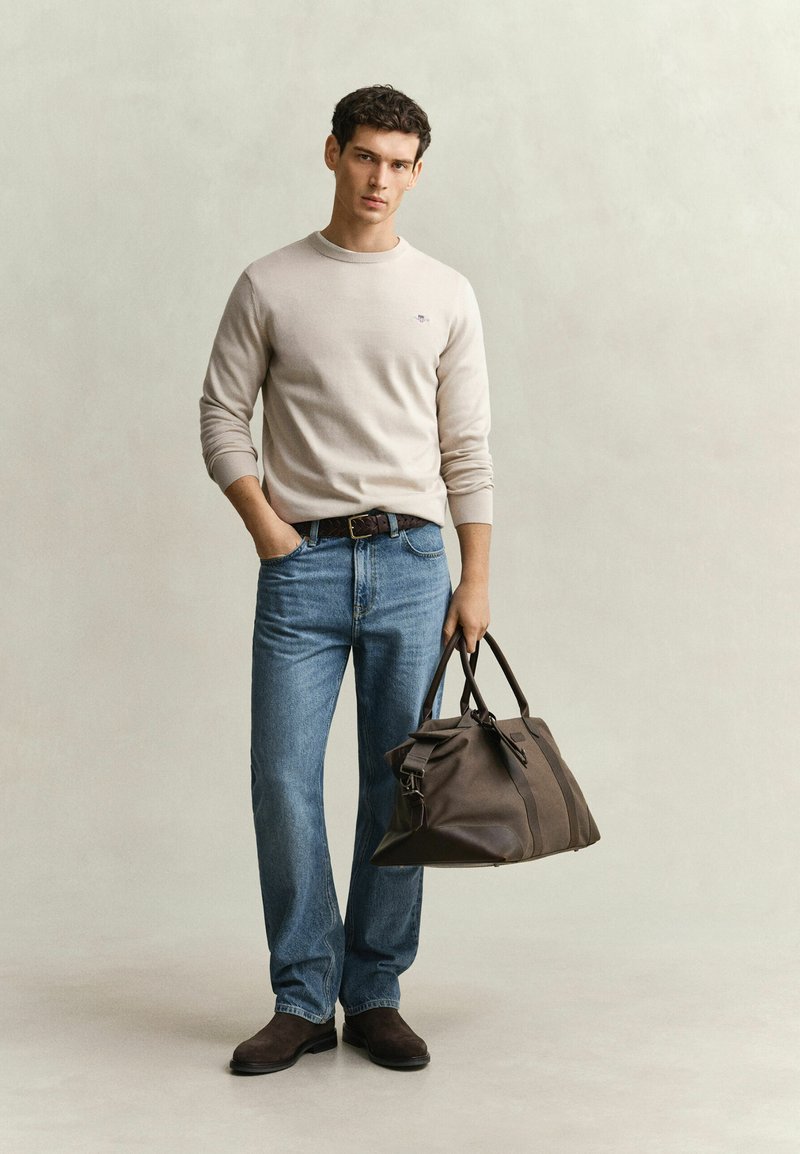 Jeune homme en pull beige et jeans bleu tenant un grand sac de voyage marron, debout devant un fond clair uni.