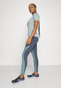 Lys blå kortermet skjorte, kombinert med marineblå og teal leggings med fargeblokkeringsdesign. Modellen har på seg marineblå treningssko.