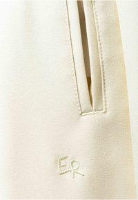 Kermanvärinen kangas lähikuvassa, jossa pystysuuntainen taskun aukko ja pieni "ER" kirjailtuna taskun alapuolelle.