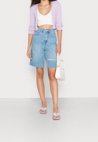 Donna che indossa un cardigan corto lilla chiaro, un top corto bianco a costine, pantaloni shorts in denim strappati a vita alta, sandali lavanda, mentre tiene in mano una piccola borsa bianca.