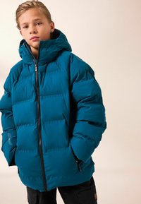 Tyrkisk pufferjakke med høj krave og hætte, der har en frontlynlås, sidelommer og en quiltet design til isolering.