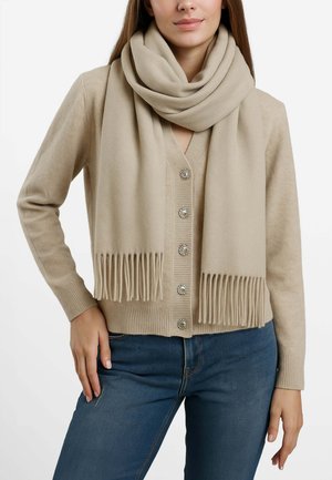 Sciarpa lavorata a maglia beige con bordi frangiati, avvolta liberamente attorno al collo, abbinata a un cardigan con bottoni coordinati e bottoni decorativi.