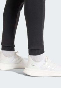 Λευκά αθλητικά παπούτσια με υφή στο πάνω μέρος, τρεις ανυψωμένες ρίγες και μαλακή σόλα, σε συνδυασμό με σκούρα αθλητική φόρμα και λευκές κάλτσες.