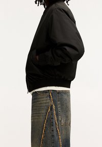 Zwarte bomberjack met elastische manchetten en zoom, met een rits, gedragen over een wit shirt, gecombineerd met versleten blauwe jeans.