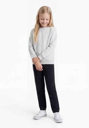 Jeune fille aux longs cheveux blonds portant un sweat-shirt gris clair, un pantalon noir et des baskets blanches, debout et souriante, les mains jointes.