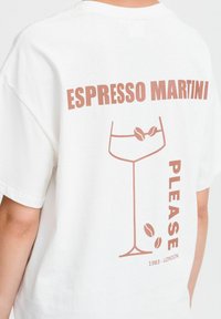 Bílý bavlněný tričko s potiskem sklenice s motivem kávových zrn a nápisem „ESPRESSO MARTINI PROSÍM“ v hnědé barvě.