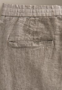Gros plan sur un pantalon en lin beige montrant la partie supérieure du dos avec une ceinture élastique et une poche passepoilée unique.