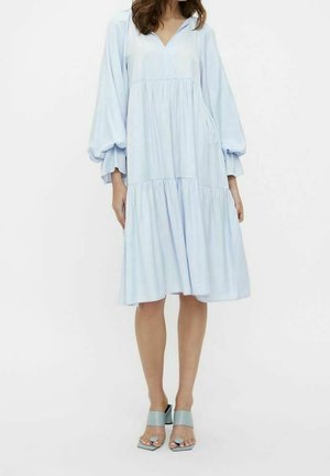 Woman wearing a light blue, loose-fitting midi dress with long puffed sleeves and open-toe block-heeled sandals, standing against a white background.