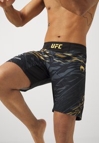UFC-brändade svarta shorts med ett strukturerat, dynamiskt mönster i nyanser av grått och guld, med en logotyp på midjebandet.