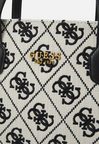 Witte canvas handtas met een zwart logopatroon, een gouden "GUESS"-embleem en zwarte leren handgrepen. Geometrisch diamantontwerp met stikselaccenten.