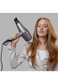 Μαλλιομούχλας με κομψό ασημένιο σχέδιο, εργονομική λαβή και μαύρη ρύθμιση. Το μοντέλο τον κρατάει μπροστά από ένα γκρι φόντο, με μακριά, κυματιστά κόκκινα μαλλιά.