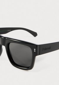 Fekete téglalap alakú Polaroid napszemüveg sötétített lencsékkel, diszkrét Polaroid márkajelzéssel a szárán és a lencse sarkán.