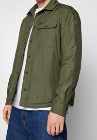 Giacca verde oliva con colletto a camicia, tasca frontale e tessuto texture. Presenta bottoni a pressione e una vestibilità casual. Abbinata a una camicia bianca.