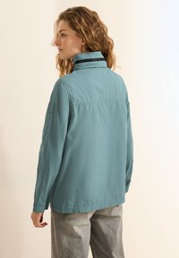 Vaaleansininen tuulitakki, jossa on korkea kaulus, slim fit -malli ja joustavat hihansuut. Takissa on hienovarainen logo vasemmassa hihassa ja matta pinta.