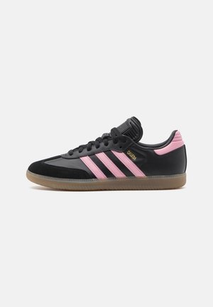 Sort og pink Adidas Samba sneaker med gummisål, tre pink striber på siden og "Samba" skrevet med guldbogstaver.
