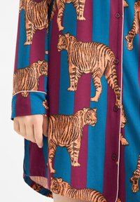 Korte mouwen shirt met een gedurfde tijgerprint in oranje tegen een blauwe en bordeauxrode gestreepte achtergrond. Knoopsluiting aan de voorkant met omgeslagen mouwen.