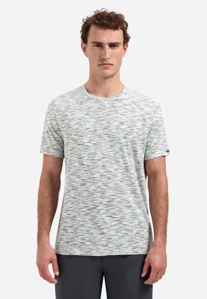 Man met krullend haar die een wit-grijs horizontaal gestreept T-shirt met korte mouwen en een donkere broek draagt, staand tegen een effen achtergrond.