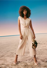 Lys beige jumpsuit med v-hals, ærmeløs design og brede bukser, fastgjort med et matchende bælte. Kombineret med strophæle og en pailletveske.