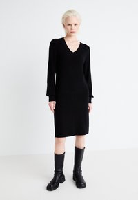 Black knit dress with a V-neckline and long sleeves, featuring a straight cut and textured fabric. Paired with knee-high black boots.