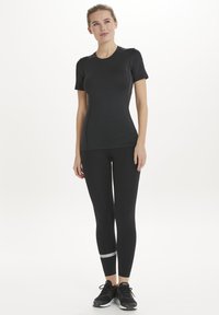 Svart atletisk topp och leggings med en slät textur; leggings har en silverdetalj vid vristen. Bärs med svarta sportskor.