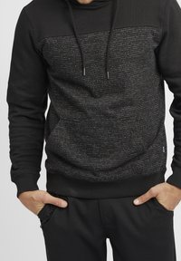 Sudadera negra con capucha y tejido texturizado que presenta rayas horizontales en gris y blanco, cordones ajustables y puños acanalados.