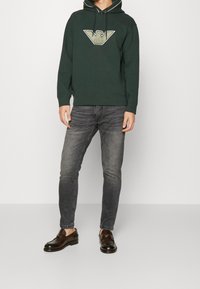 Grön hoodie med en emblemet i guld och beige, kombinerad med mörkgrå jeans och svarta loafers. Hoodien har en dragsko och ribbade ärmar.