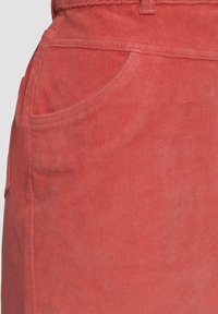 Tissu en velours côtelé corail, présentant une finition texturée douce, avec une poche latérale et des détails de couture visibles le long des bords.