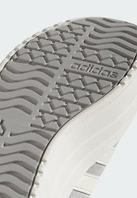 Adidasova potplat cipela s teksturiranim siva gumenom podlogom s uzorakom u obliku cik-caka i urezanim logotipom branda. Bočni pogled prikazuje bijeli gornji dio.