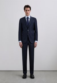 Traje azul marino con chaqueta estructurada que cuenta con dos bolsillos frontales y cierre de botones, combinado con una camisa de vestir azul claro y corbata oscura. Zapatos negros completan el conjunto.