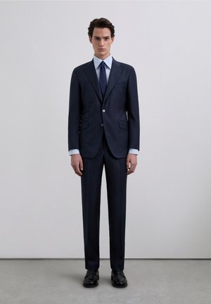 Traje azul marino con chaqueta estructurada que cuenta con dos bolsillos frontales y cierre de botones, combinado con una camisa de vestir azul claro y corbata oscura. Zapatos negros completan el conjunto.